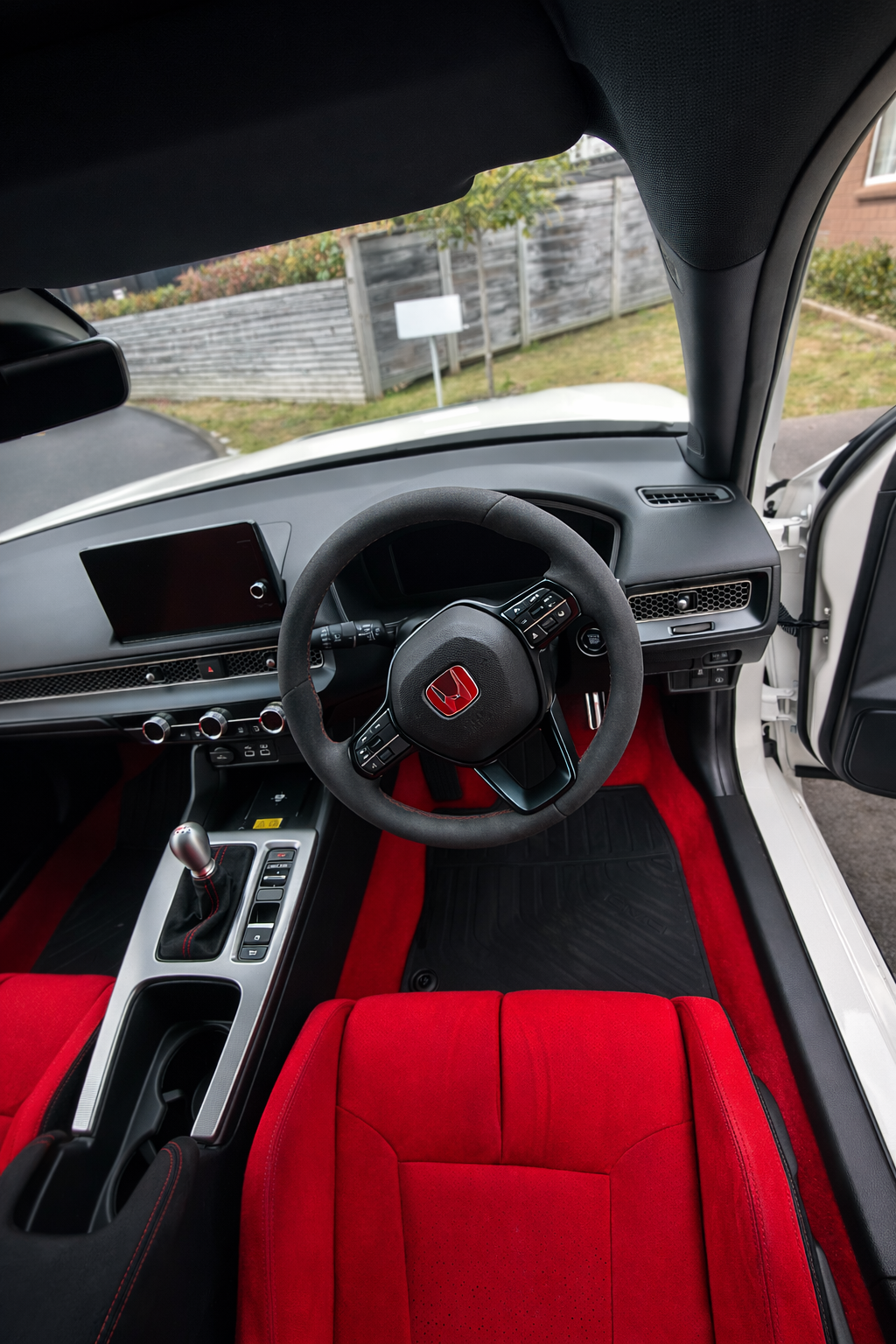 Car interior with red seats and steering wheel, visible outside is a residential area.